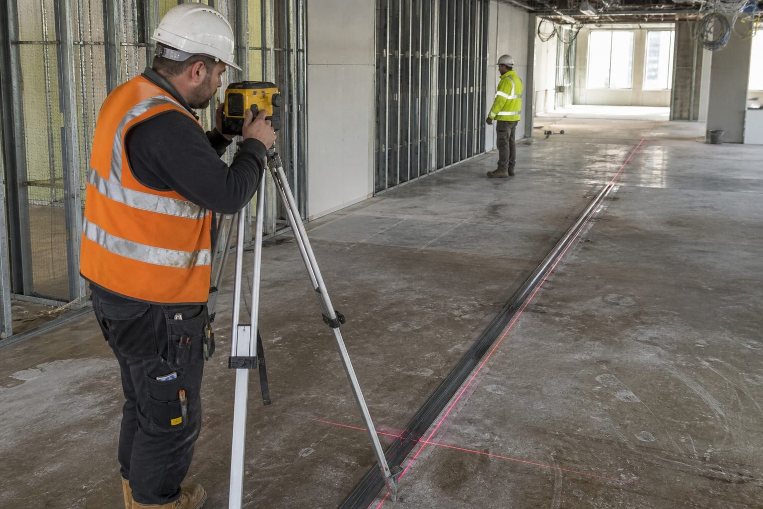 Setting out partition floor track with a laser level on a UK commercial fit-out, operatives in PPE.